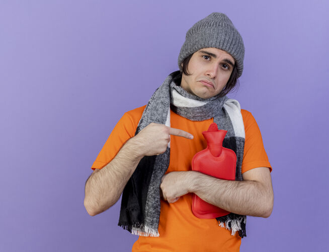 围巾一个戴着冬天帽子 戴着围巾的悲伤的生病的年轻人拥抱着 指着孤立在紫色背景上的热水袋热帽子年轻