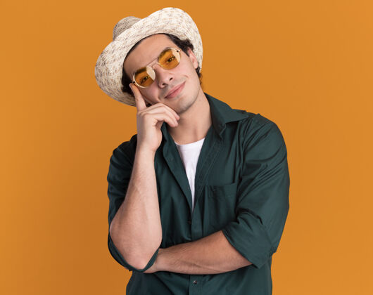 快乐身穿绿色衬衫 戴着夏季帽子 戴着眼镜 脸上带着微笑站在橙色的墙上的快乐的年轻人年轻眼镜戴着