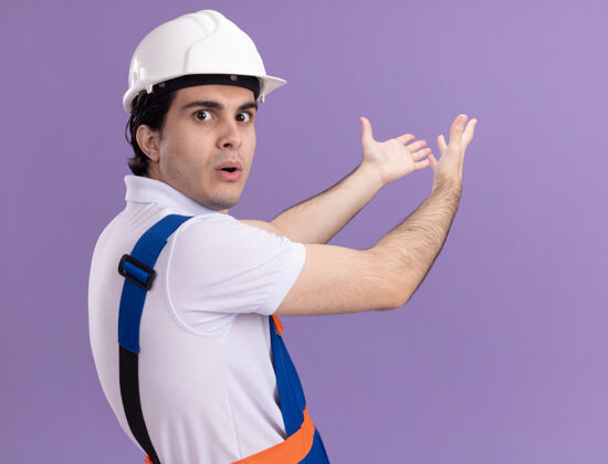 介绍年轻的建筑工人穿着建筑制服 戴着安全帽 看上去很困惑 他伸出双臂站在紫色的墙上建筑困惑建设者