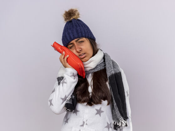 围巾悲伤的生病的年轻女孩戴着冬天的帽子戴着围巾把热水袋放在脸颊上隔离在白色的背景上脸颊女孩包
