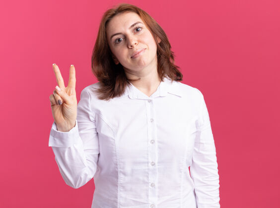 Vsign身穿白衬衫的年轻女子面带微笑地看着前面 粉红色的墙上立着v字标志衬衫女人表演