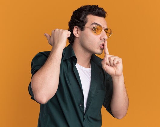 做着身穿绿色衬衫 戴着眼镜的年轻人站在橙色的墙上 手指放在嘴唇上做着沉默的手势沉默眼镜放在一边