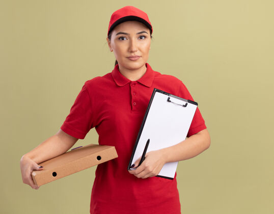 披萨身穿红色制服 头戴鸭舌帽的年轻女送货员拿着披萨盒和剪贴板 手里拿着白纸看着前面 严肃的脸站在绿色的墙上拿着盒子送货
