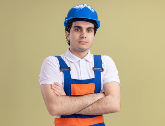 头盔年轻的建筑工人穿着建筑制服 戴着安全帽 双手交叉着站在绿色的墙上 自信地看着前方站立年轻建筑