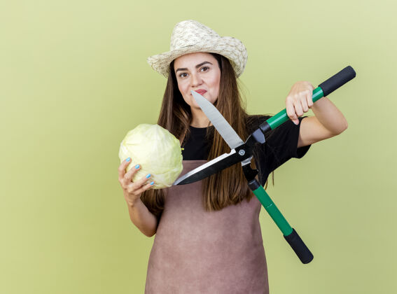 卷心菜高兴美丽的园丁女孩穿着制服 戴着园艺帽 试图削减卷心菜与剪子孤立在橄榄绿的背景试试帽子切割