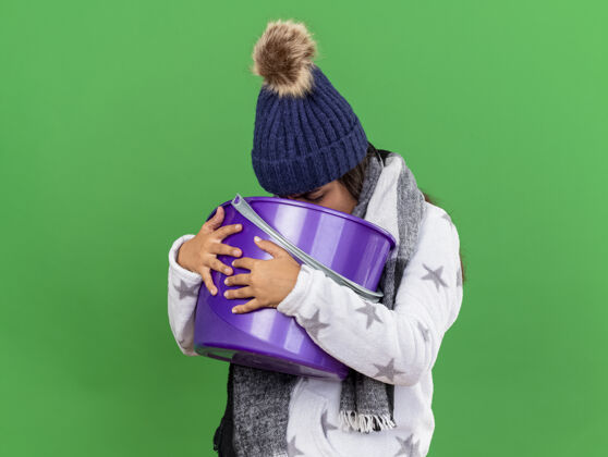 围巾年轻的生病女孩戴着冬天的帽子 戴着围巾 手里拿着塑料桶 把呕吐物放在绿色的背景上水桶生病呕吐