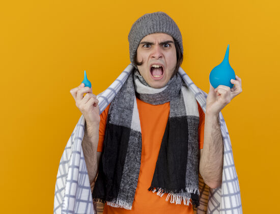 年轻愤怒的年轻病人戴着冬天的帽子 围巾裹着格子布 拿着隔离在橙色背景上的灌肠剂抱着围巾格子