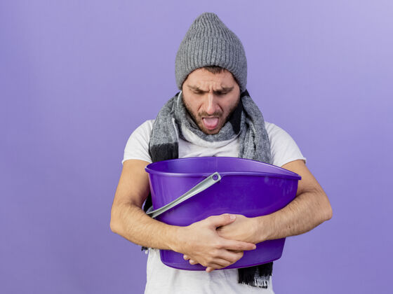 呕吐一个戴着带围巾的冬帽 拿着塑料桶 吐在紫色背景上的年轻人塑料年轻持有
