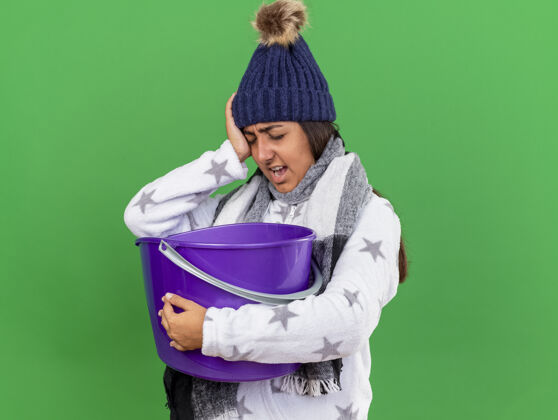 不愉快戴着冬天的帽子戴着围巾拿着塑料桶手放在眼睛上的未被释放的生病的年轻女孩在绿色背景上孤立年轻持有水桶