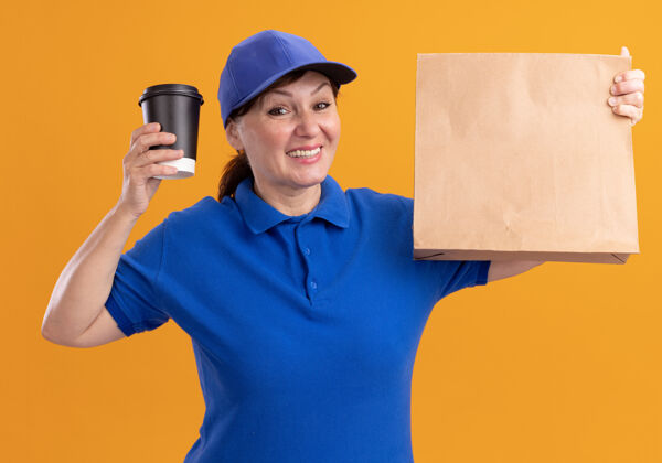 送货中年女送货员身穿蓝色制服 戴着帽子 手里拿着纸包 手里拿着咖啡杯 看着前面 站在橘色的墙上开心地微笑着包装表演女人