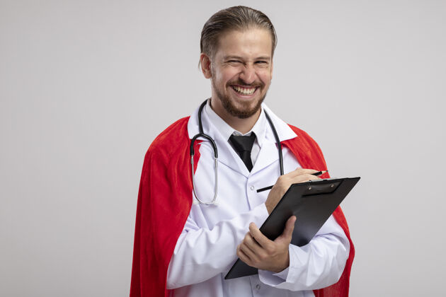 铅笔微笑着的年轻超级英雄 穿着医用长袍 听诊器拿着剪贴板 铅笔隔离在白色背景上剪贴板超级英雄听诊器