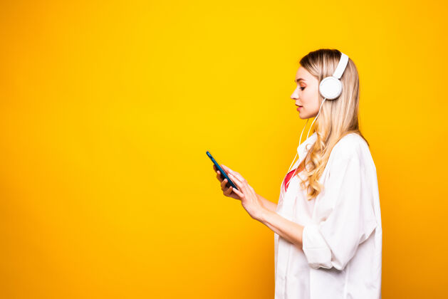 肖像一个戴着耳机和手机隔着黄墙听音乐的快乐年轻女子的画像手手机电话