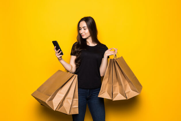 购物漂亮的年轻女子拿着购物袋在黄色的墙上用她的智能手机购物购物狂购物时尚夏天携带快乐