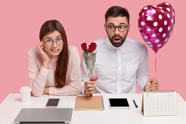 年轻人一对年轻夫妇坐在书桌旁 一个男人手里拿着一束玫瑰花和心形气球平板电脑男性女人