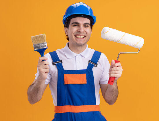 站着年轻的建筑工人身穿建筑制服 戴着安全帽 拿着油漆刷和压路机 站在橙色的墙上 面带微笑地看着前方安全建筑建筑工人