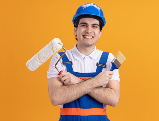 抱着年轻的建筑工人身穿建筑制服 戴着安全帽 拿着油漆刷和滚筒 站在橙色的墙上 面带微笑地看着前方头盔建筑工人微笑