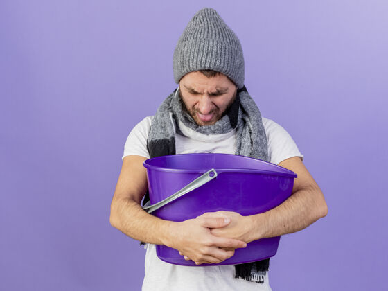 围巾低着头 戴着冬天的帽子 戴着围巾 拿着塑料桶 吐在紫色的背景上持有不愉快年轻