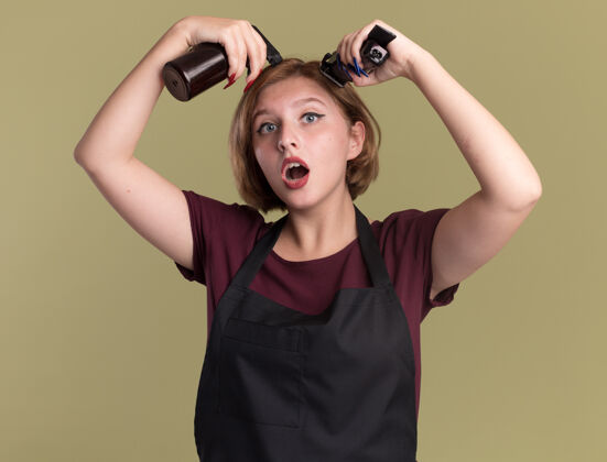 围裙年轻漂亮的女理发师站在绿色的墙上 拿着喷雾瓶和修剪机 围着围裙剪头发机器年轻女人