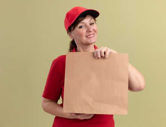 站着身穿红色制服 头戴鸭舌帽的中年女送货员拿着纸包望着前面 微笑着自信地站在绿色的墙上微笑抱着制服