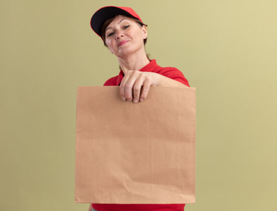 抱着身穿红色制服 头戴鸭舌帽的中年女送货员拿着纸包望着前面 微笑着自信地站在绿色的墙上纸女人中年