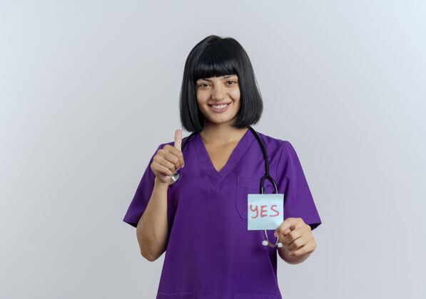 制服微笑的年轻黑发女医生穿着制服 手持听诊器 举着“是”字条 竖起大拇指笔记是持有