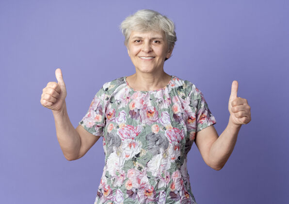 拇指微笑的老妇人竖起大拇指 两只手孤立在紫色的墙上手老人老人