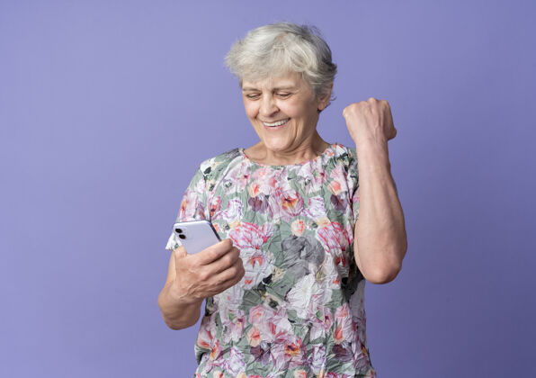 老人微笑的老妇人举起拳头看着紫色墙上孤立的电话电话拳头微笑