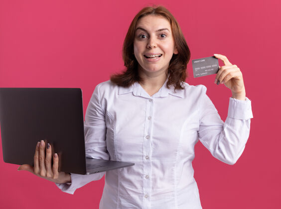 年轻穿着白衬衫的年轻女子拿着笔记本电脑 看着前面的信用卡 站在粉色的墙上 她笑得很开心信用微笑笔记本电脑