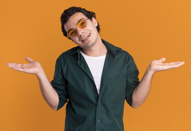 男人身穿绿色衬衫 戴着眼镜的年轻人面带微笑地站在橙色的墙上 向两边伸出双臂站着眼镜穿上