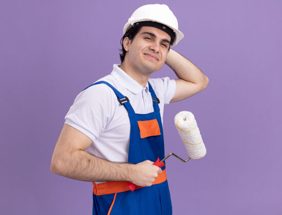 年轻人年轻的建筑工人身穿建筑制服 戴着安全帽 手里拿着油漆辊 站在紫色的墙上开心地看着一边滚轴制服抱着
