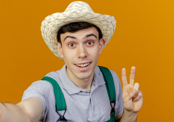 橙色快乐的年轻男园丁戴着园艺帽 打着胜利手势 假装拿着相机手喜悦复制