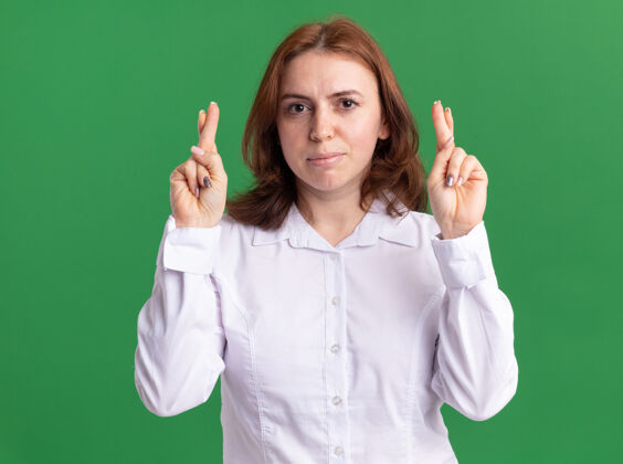 女人穿白衬衫的年轻女子站在绿色的墙上 严肃地看着前面 手指交叉着脸手指年轻
