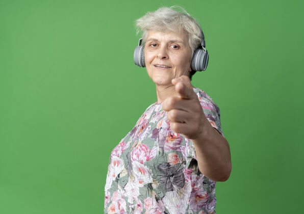 耳机戴着耳机微笑的老妇人看着绿墙上孤立的点女人老人老的