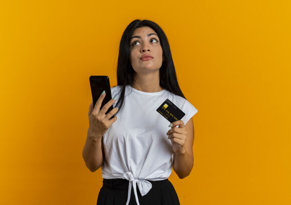 橙色体贴的年轻白人女孩拿着电话和信用卡看着身边壁板电话复制