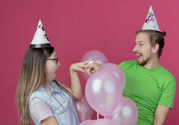 氦一对戴着派对帽的年轻夫妇惊讶地看着对方 指着站在粉红色墙上的氦气球每个站情侣