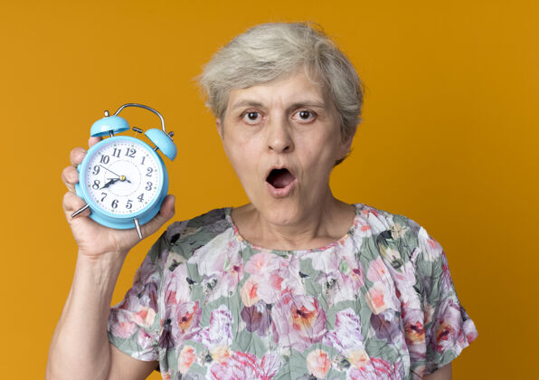 老人惊讶的老妇人把闹钟隔离在橙色的墙上时钟惊喜女人