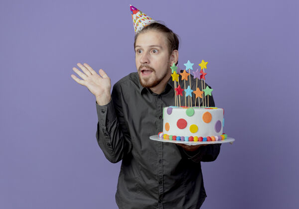 举行惊讶的帅哥戴着生日帽拿着蛋糕举手看着紫色墙上孤立的一面帽子蛋糕男人