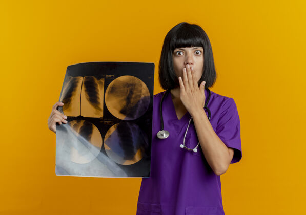 嘴巴震惊的年轻黑发女医生穿着制服用听诊器把手放在嘴里拿着x光结果橙色结果拿着