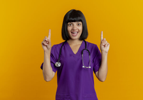 惊喜惊讶的年轻黑发女医生穿着制服 用听诊器双手指向上橙色手太空