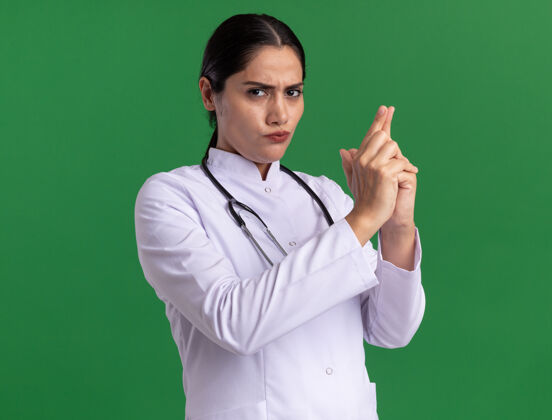 女人年轻的女医生穿着医用外套 拿着听诊器 严肃地看着前面 用手指站在绿色的墙上做着一个手势听诊器做医生