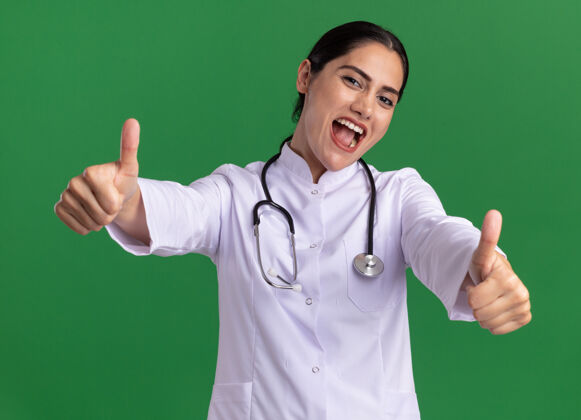 快乐年轻的女医生 身穿医用外套 脖子上戴着听诊器 脸上挂着幸福的笑容 站在绿色的墙上 高兴地竖起大拇指脸围着欢呼