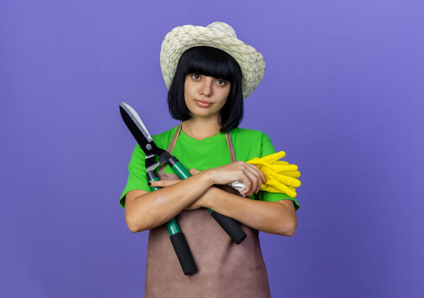 剪子自信的年轻女园丁穿着制服戴着园艺帽交叉双臂拿着剪刀和手套交叉空间女