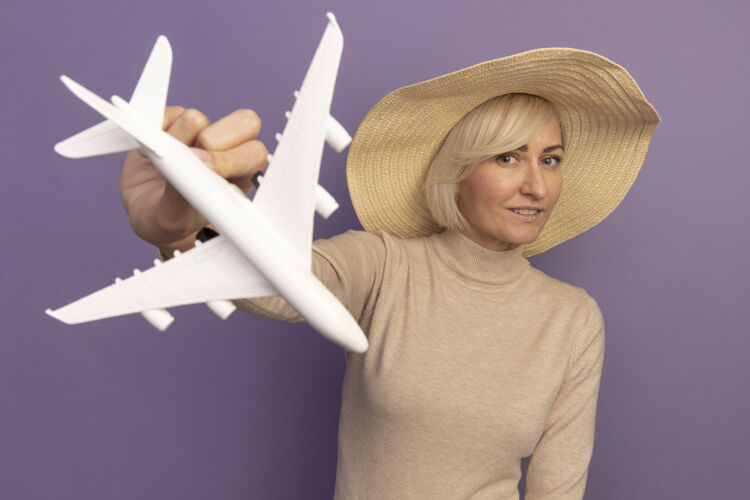 请戴着沙滩帽的漂亮金发斯拉夫女人拿着紫色的飞机模型时尚帽子时尚