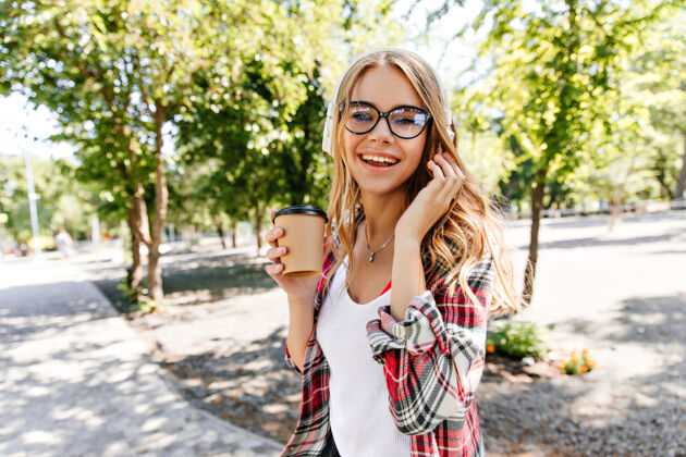 休闲迷人的戴眼镜的年轻女子拿着一杯咖啡在大自然中漫步微笑的金发女孩在夏日公园里漫步女人放松积极