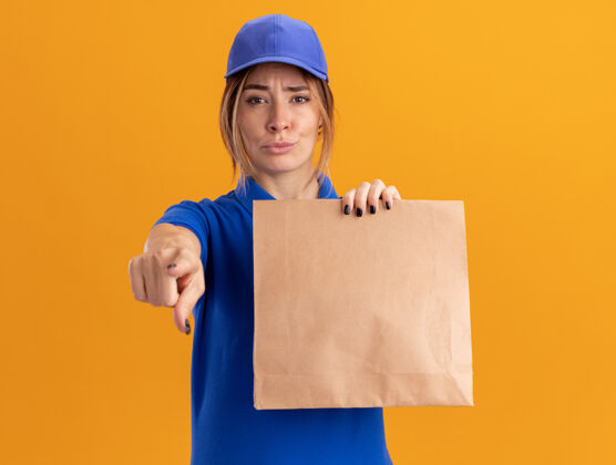 漂亮穿着制服的年轻漂亮的送货女孩拿着纸包 指着橙色的相机点姿势包装