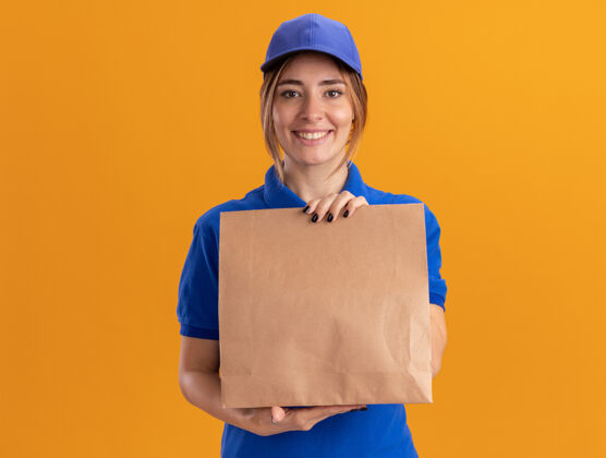 包装微笑着的年轻漂亮的送货女孩 穿着制服 手里拿着橘黄色的纸包制服摆姿势年轻