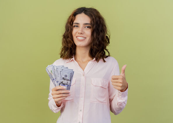 微笑微笑的年轻白种女人拿着钱 竖起大拇指孤立地放在绿色背景上 留着复印空间年轻女人拇指