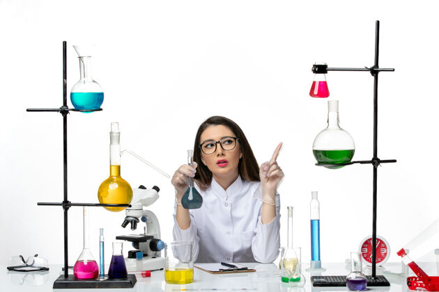 套装正面图：穿着白色医疗服的女化学家拿着装有蓝色溶液的烧瓶 放在白色办公桌上 实验室科学病毒柯维德大流行正面玻璃科学