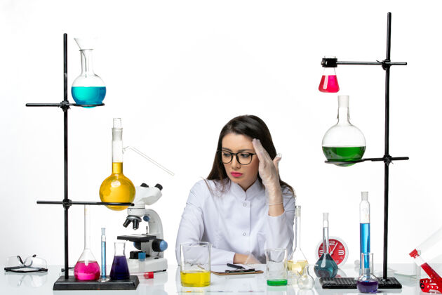 大流行前视图穿着白色医疗服的女化学家坐在浅白背景上 拿着不同的溶液科学大流行病毒冠状病毒实验室人解决方案成人