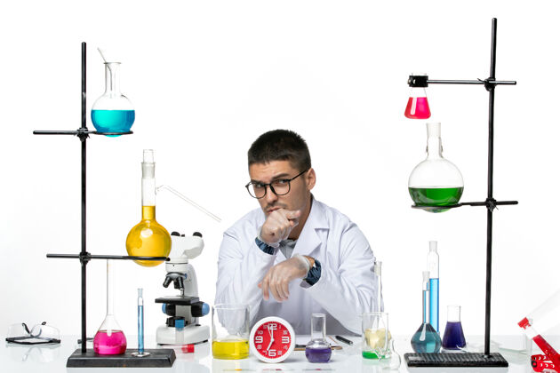 视图正面图身着白色医疗服的男性化学家坐在浅白背景上 用不同的溶液研究病毒疾病科学实验室的科维德前面病毒不同